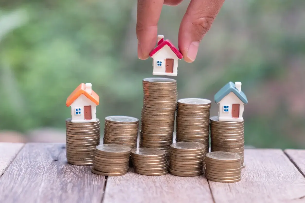 Photo de 3 maisons miniatures sur des piles de pièces, concept d'estimation immobilière. Utilisée dans l'article Building Partners sur la façon d'estimer son bien à Paris 16 en 2026. Crédits iStock : Kanjana Jorruang. Référence de la photo : 1150292172.